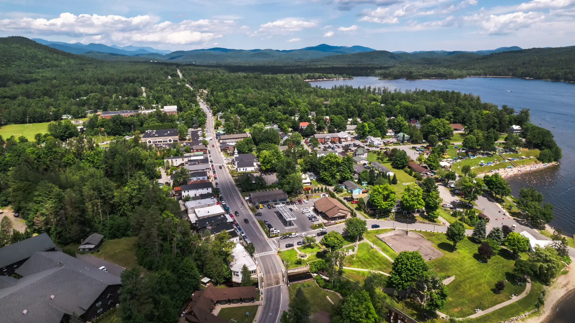 An aerial view of the town of Schroon Lake, featuring a main road lined with buildings, a beach, and a calm lake on the right