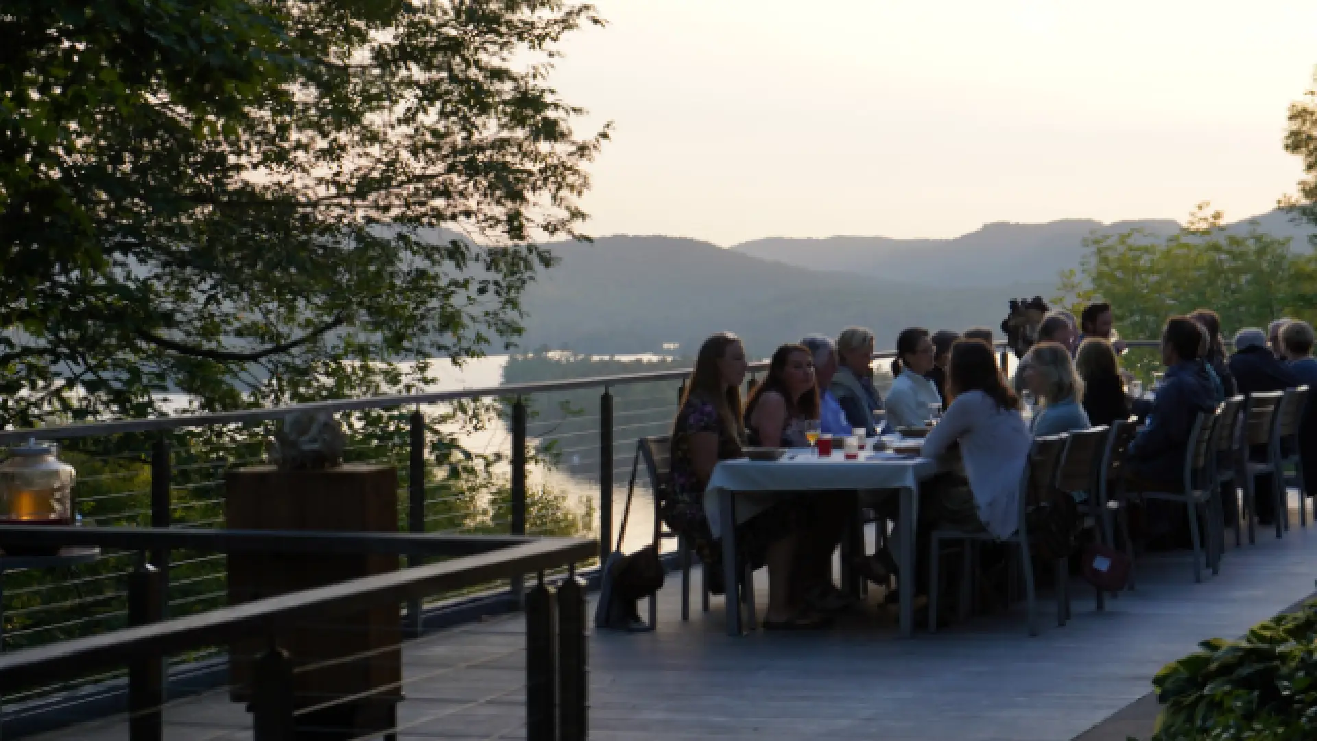 A large group of people sit at a long table set for dining on a deck overlooking a body of water with the Adirondack mountains in the background.