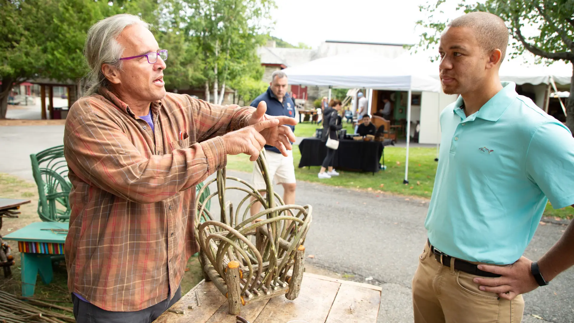 Two men talking, one man is an artisan making furniture from twigs and thin branches and the other man visiting the vender and listening