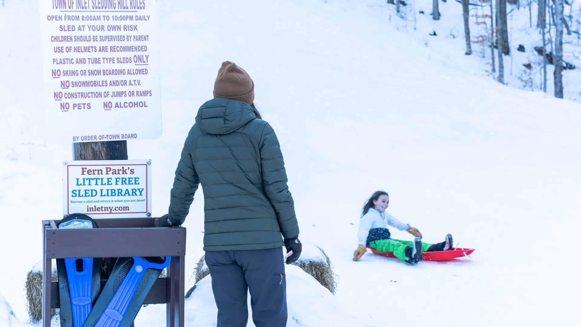A kid sledding down a hill as her parent watches.