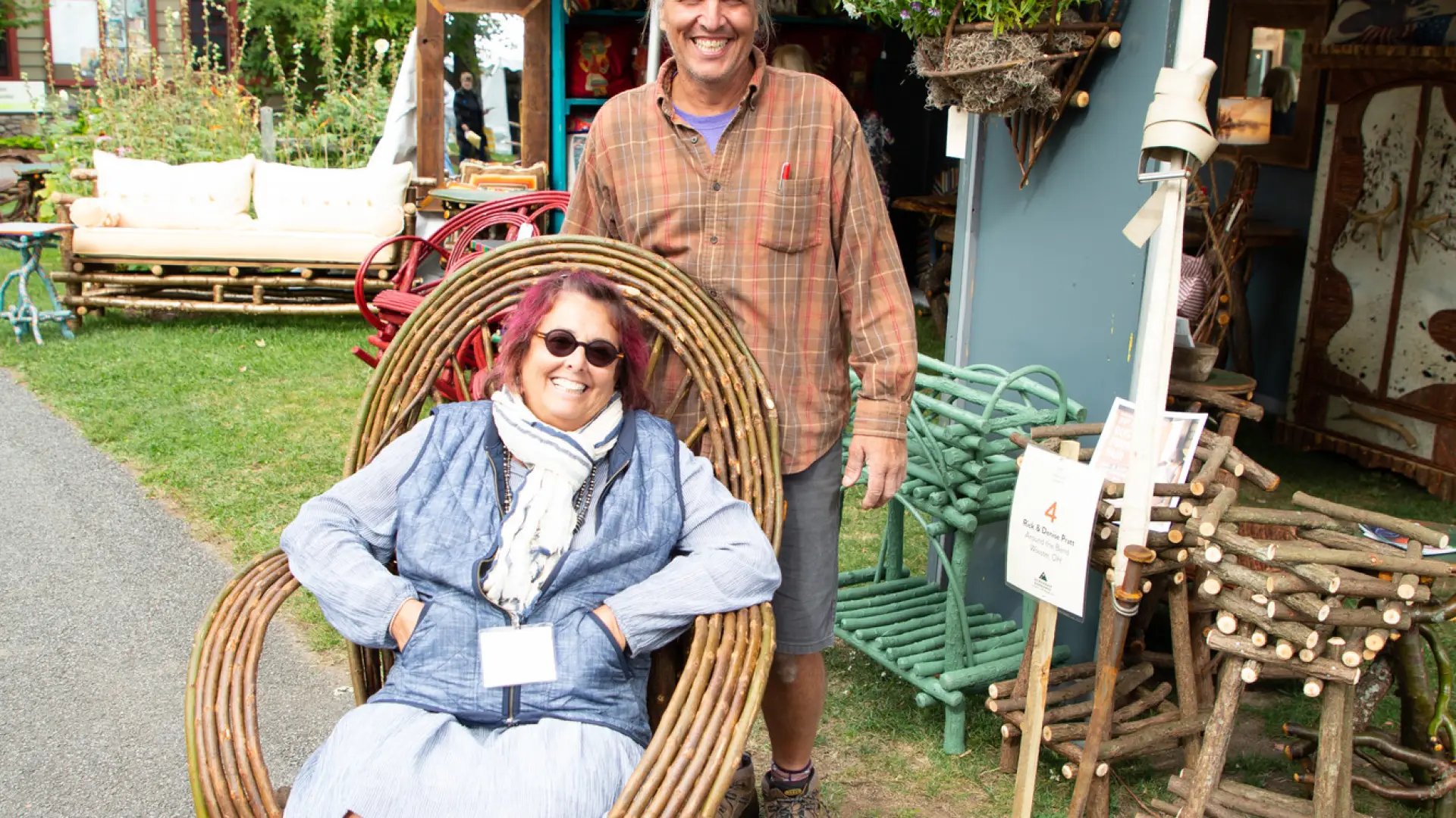 Woman sitting in a rocking chair made of thin branches with a mans standing behind her, both smiling for the photo