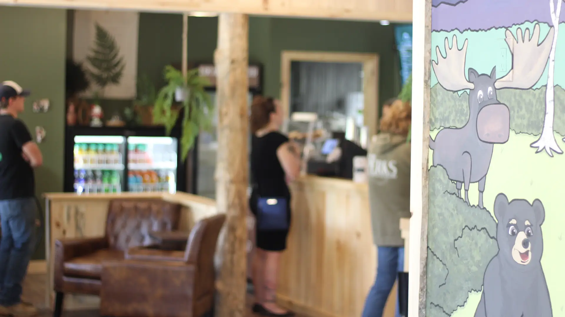 A view of the mural on the wall depicting a moose, bear and other wilderness scene with a set of leather chairs in a cozy corner, the drink fridge and people lined up to get their order in.