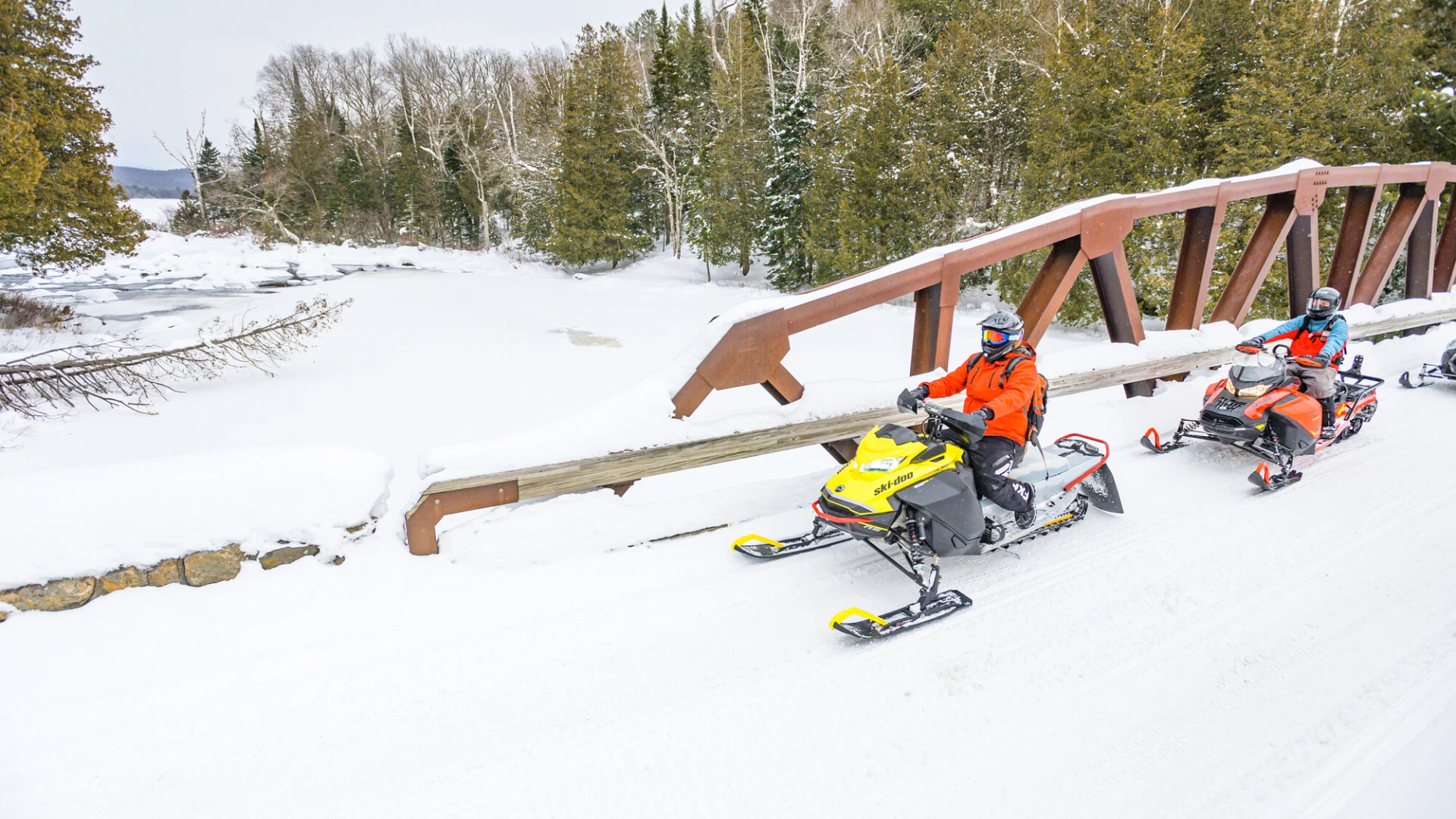 Two people in orange jackets ride snowmobiles on a snowy trail. They cross a wooden bridge amid a wintry landscape with evergreen trees.