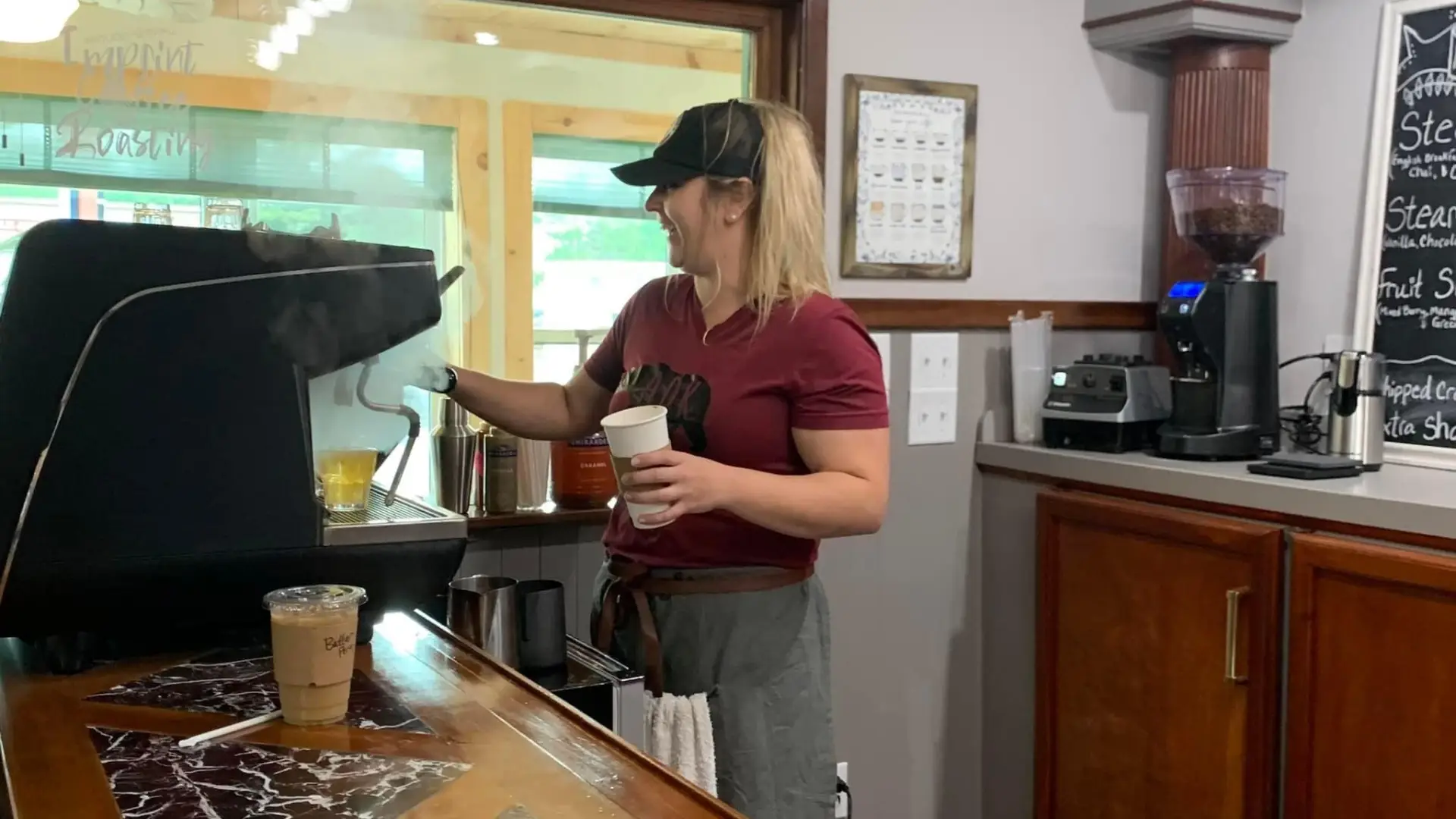 A happy blond woman operates the espresso machine while steam rises from the preparation.