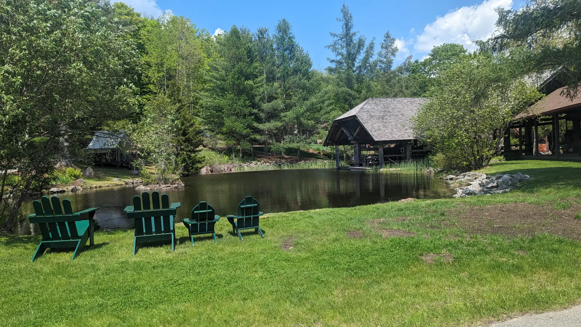 A small pond surrounded with grasses, some Adirondack style chairs for adults and kids, and two large gazebos