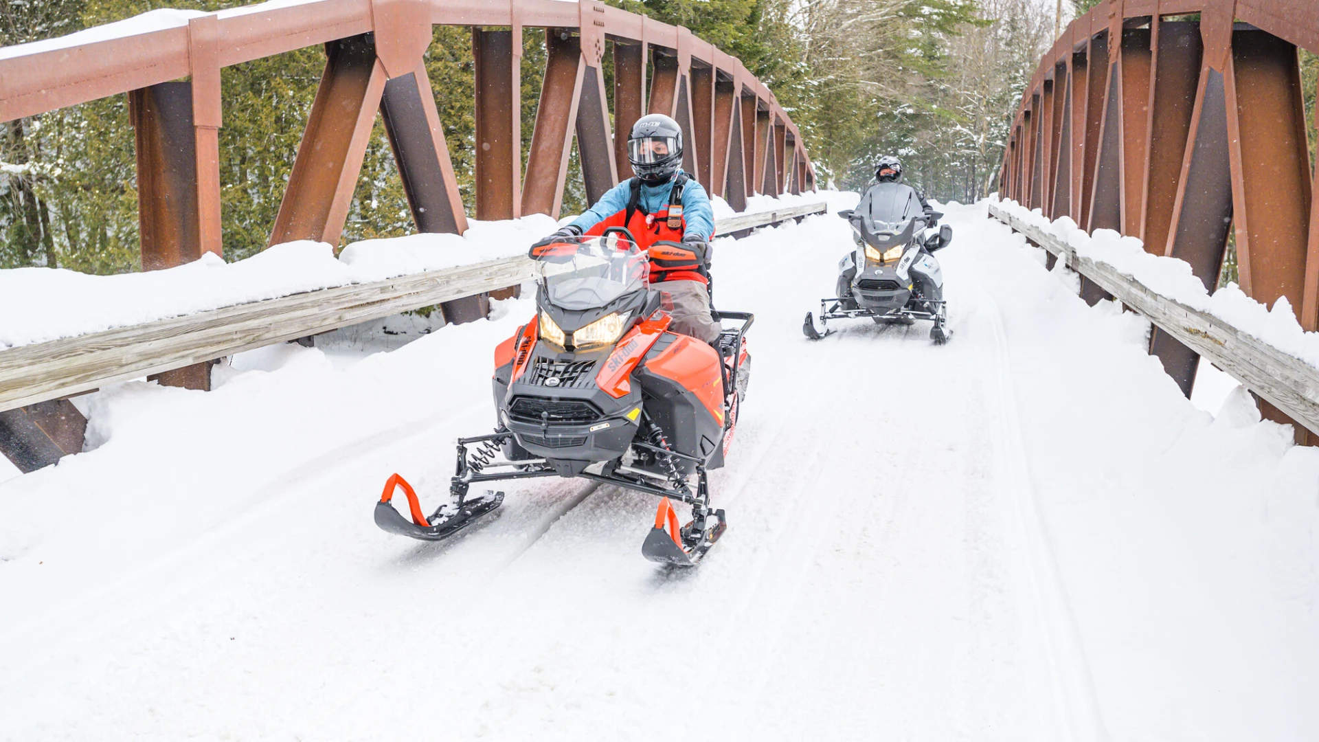 Two people ride snowmobiles across a snowy, rustic bridge. 