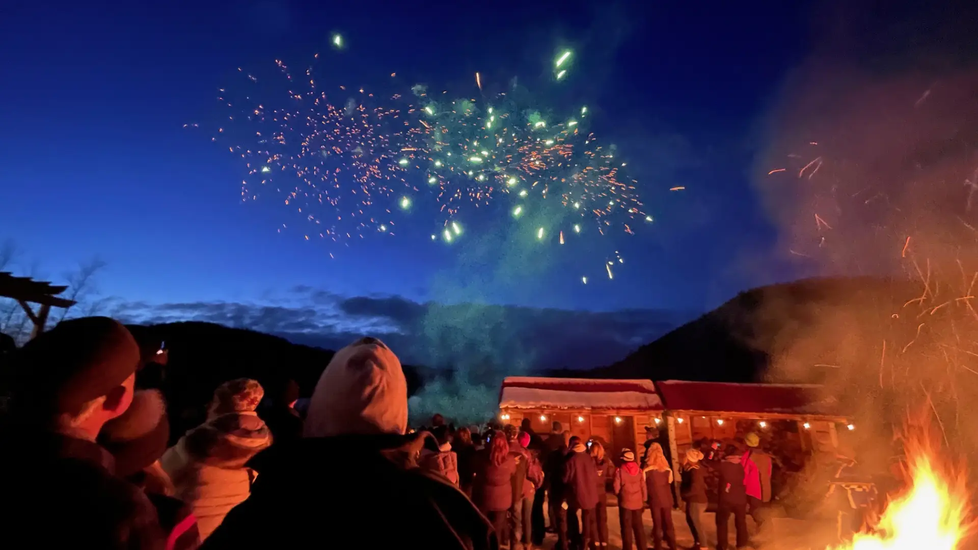 Fireworks bursting in the night sky as people watch standing around the bonfire