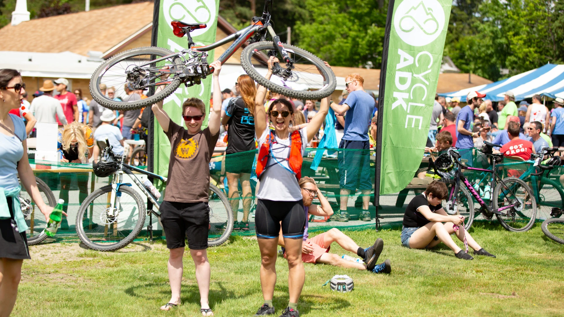 Two people joyfully lift a bicycle above their heads at an outdoor cycling event. Green "Cycle ADK" banners are in the background.