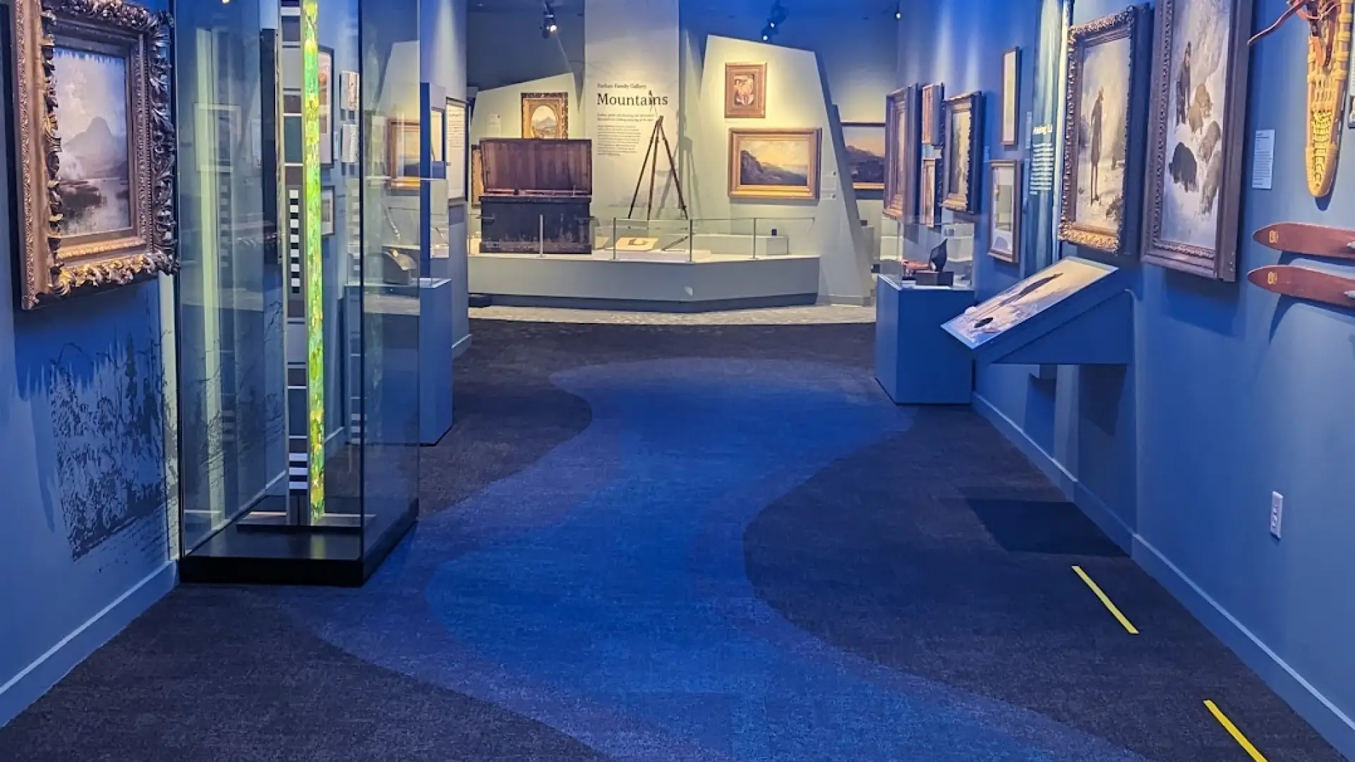 Art pieces on display in a long hallway with blue walls, a blue carpet with a winding pathway design and blue light fixture that resembles waves