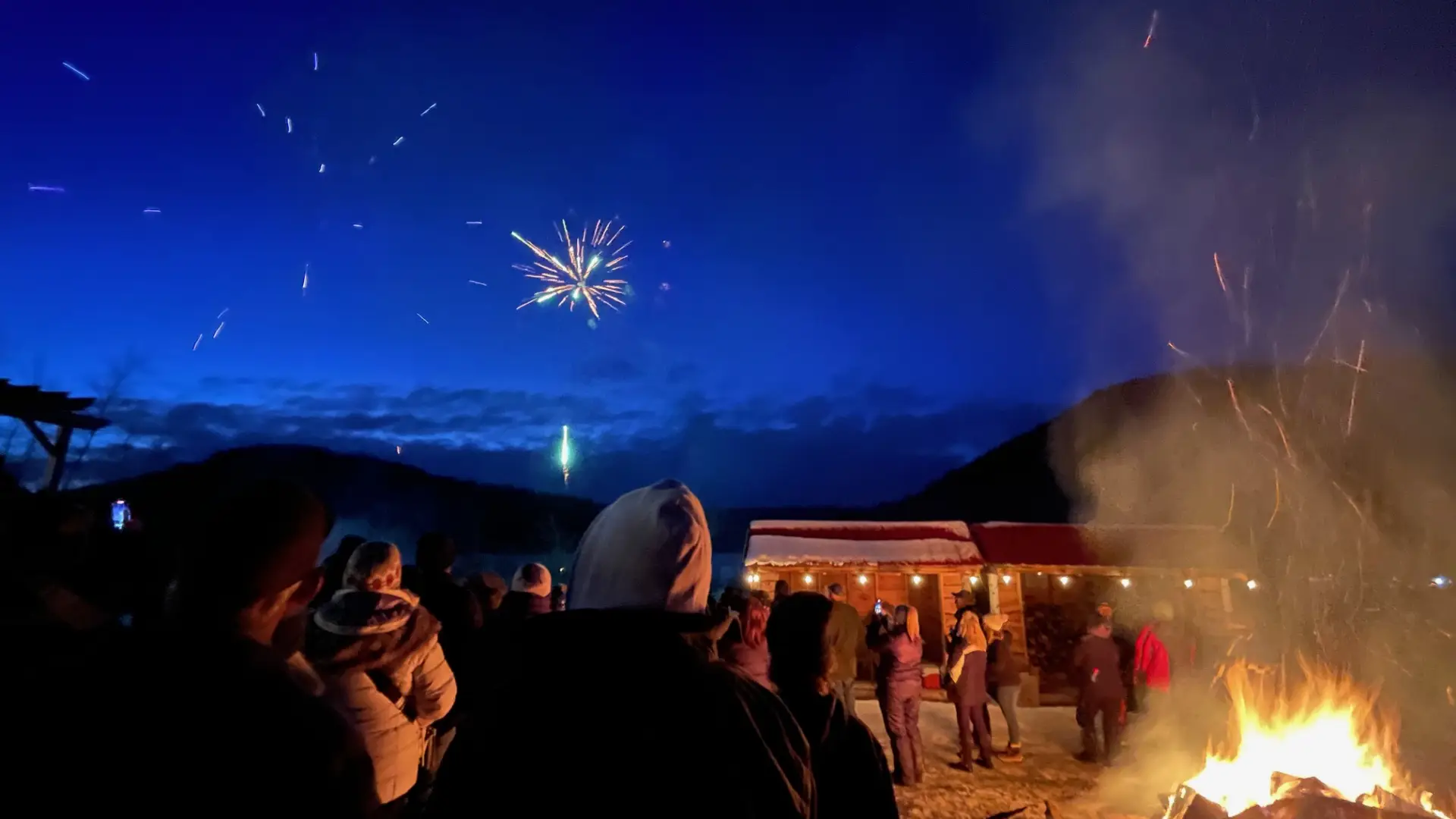 Fireworks bursting in the night sky as people watch standing around the bonfire