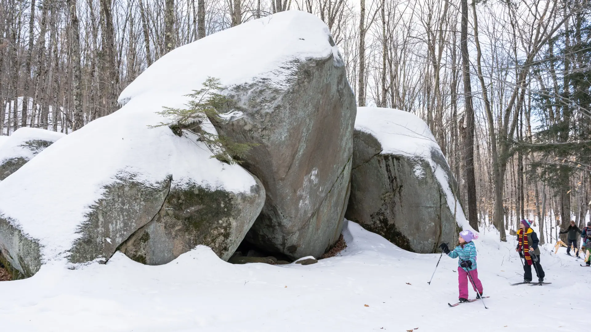 Two kids cross-country ski past tall boulders