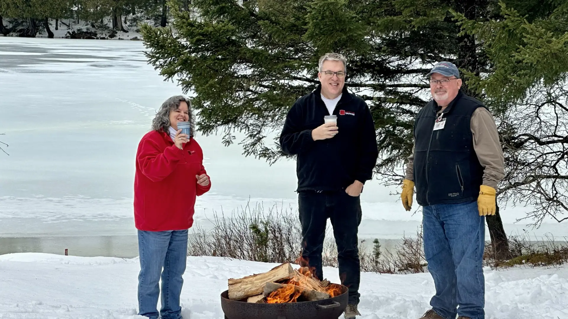 Three people standing around a campfire outside in the snow drinking warm beverages