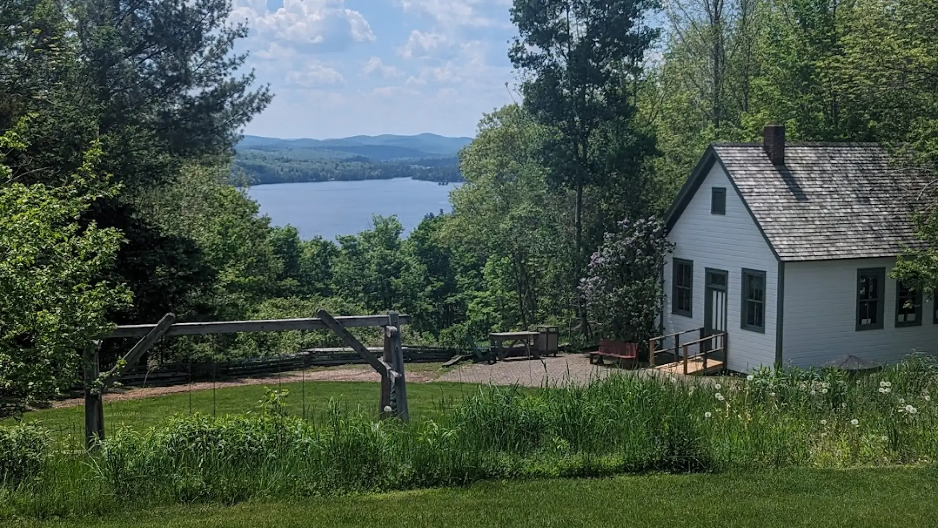 Blue Mountain Lake under billowing white clouds across a lush green lawn in front of a quaint white cabin.
