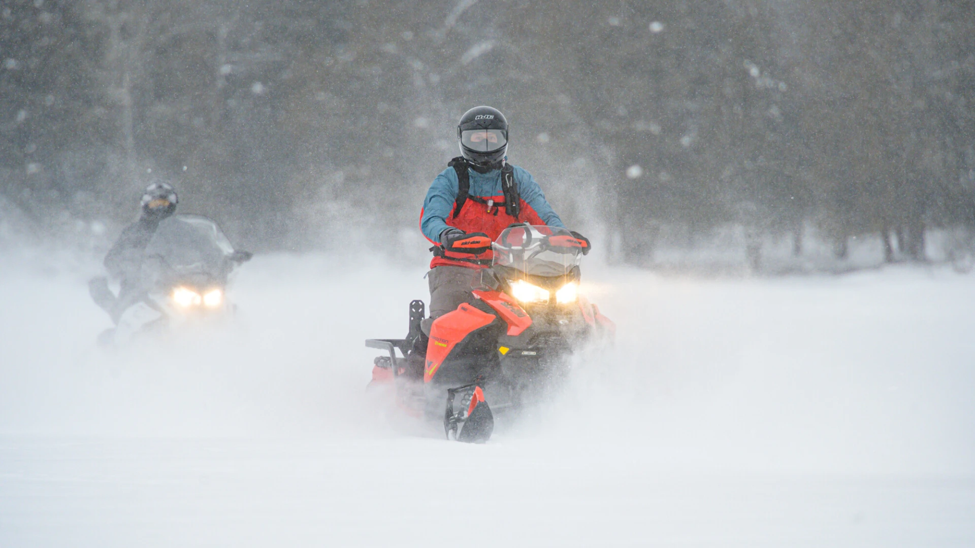 Two people ride snowmobiles through a snowy landscape. 