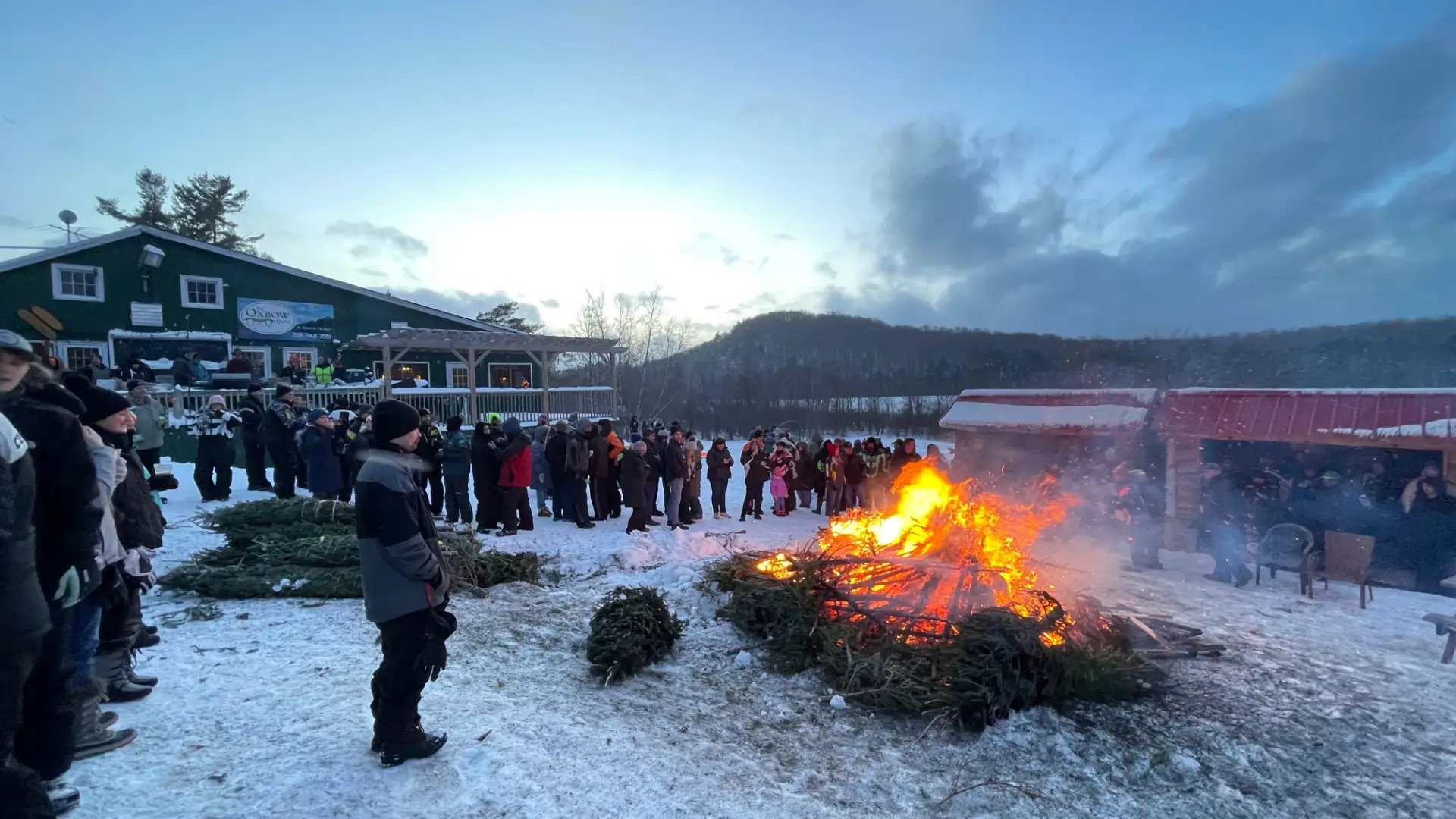 the tree bon fire with a crowd of people circled around with the Oxbow Inn in the background