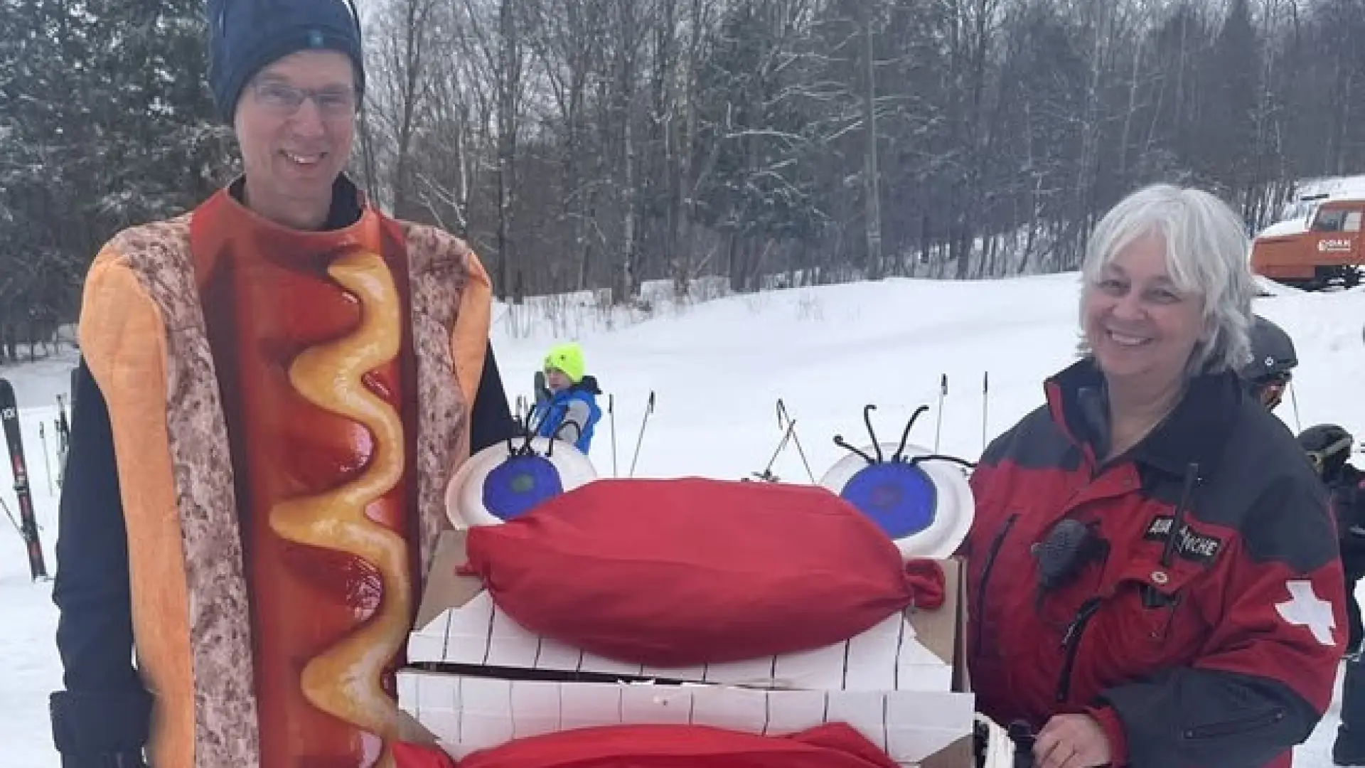 A mad dressed up on a hot dog costume as he and a women hold up a silly cardboard sled