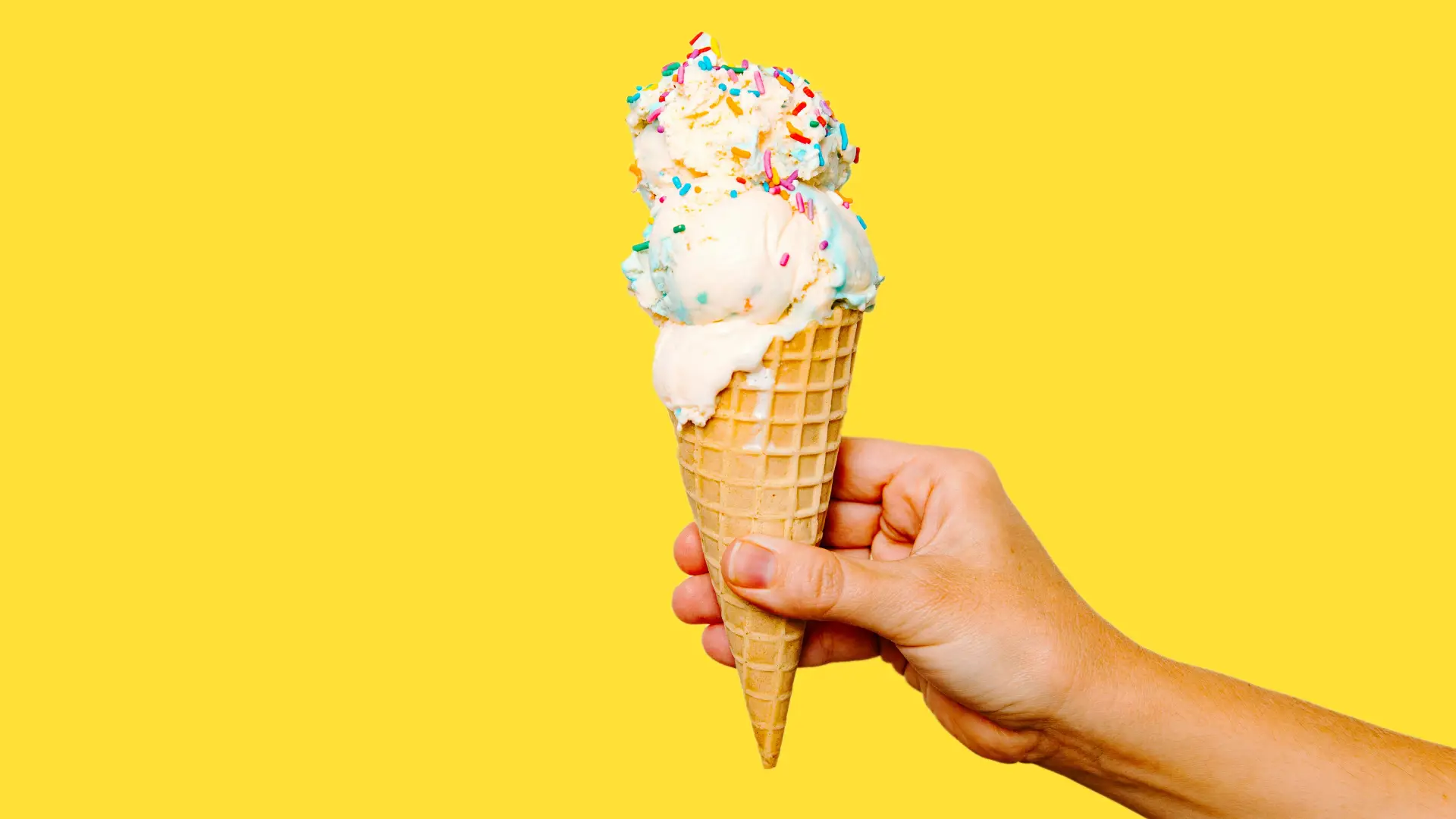 A hand holds a double-scoop ice cream cone topped with rainbow sprinkles against a solid, bright yellow background.