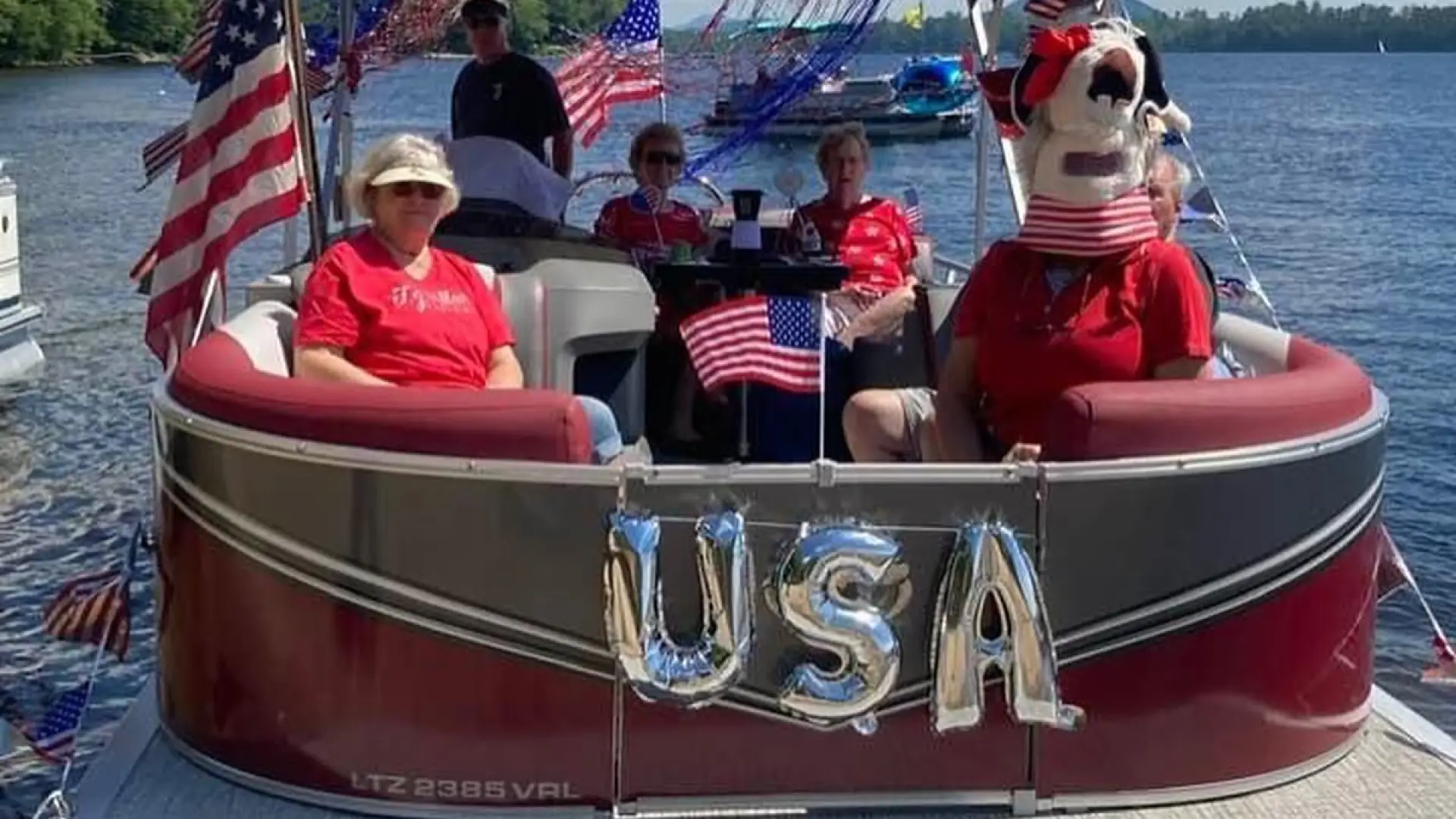 A pontoon boat decorated for the Fourth of July with American flags, streamers, and silver "USA" balloons. Several people are on board, one wearing a dog head.
