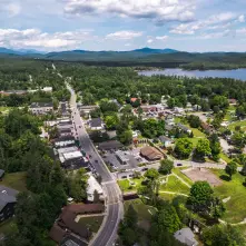 An aerial view of the town of Schroon Lake, featuring a main road lined with buildings, a beach, and a calm lake on the right