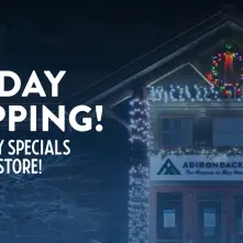 Text reads: Holiday Shopping, Shop Holiday Specials in the ADKX Store!  Image of the Adirondack Experience Visitor Center at night with bright white LED lights and a colorful wreath.