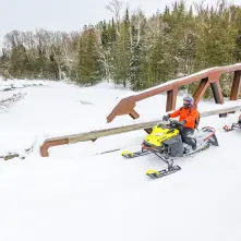 Two people in orange jackets ride snowmobiles on a snowy trail. They cross a wooden bridge amid a wintry landscape with evergreen trees.