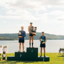 Three male runners stand on a three-tiered winner's podium by a lake, each holding a carved bear trophy.