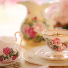 A porcelain tea set with a dark pink rose motif sits on linen table cloth covered surface