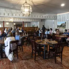 A grand dining room with chandeliers and white paneled walls filled with large wooden dining tables.