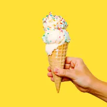 A hand holds a double-scoop ice cream cone topped with rainbow sprinkles against a solid, bright yellow background.