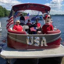 A pontoon boat decorated for the Fourth of July with American flags, streamers, and silver "USA" balloons. Several people are on board, one wearing a dog head.
