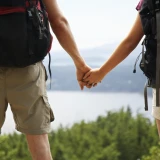 Two people hold hands with backpacks on and looking at a view with trees and a body of water.