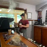 A happy blond woman operates the espresso machine while steam rises from the preparation.