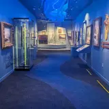 Art pieces on display in a long hallway with blue walls, a blue carpet with a winding pathway design and blue light fixture that resembles waves