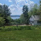 Blue Mountain Lake under billowing white clouds across a lush green lawn in front of a quaint white cabin.