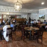 A grand dining room with simple chandeliers and white paneled walls filled with large wooden dining tables.