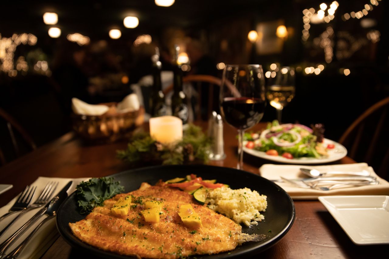 A plate of trout and a salad with a glass of wine on a table in a low-lit lodge restaurant. 