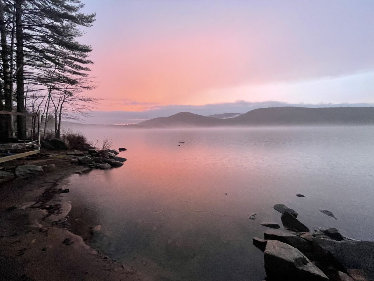 A calm lake is lit pink by a sunset with low mountains on the far shoreline.