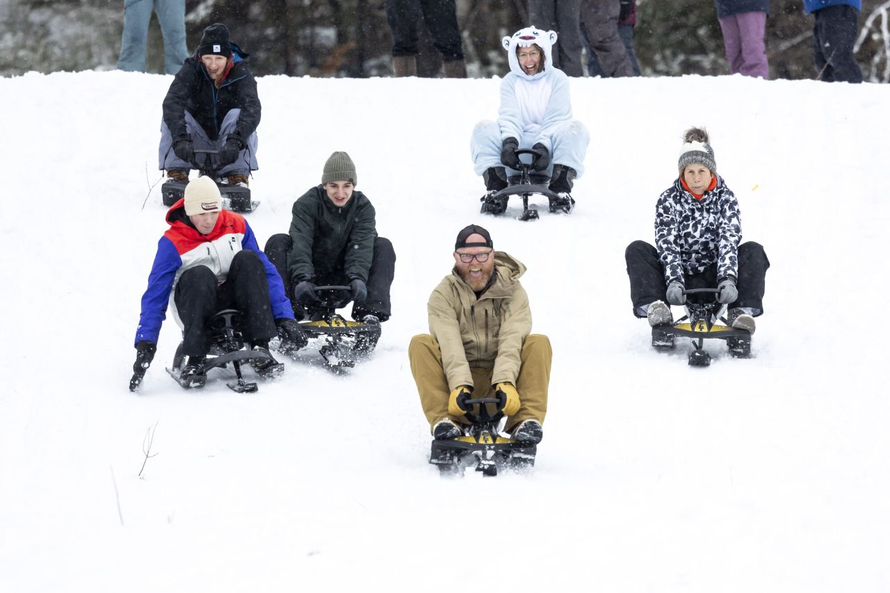 A group of adults laugh as they sled down a snowy hill. 
