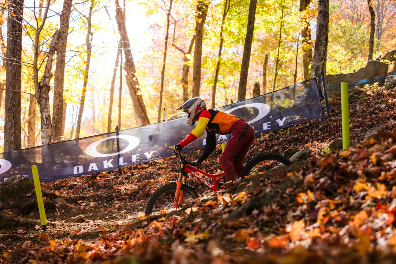 UCI MTB World Cup competitor races mountain bike down forested track with Oakley banner in background