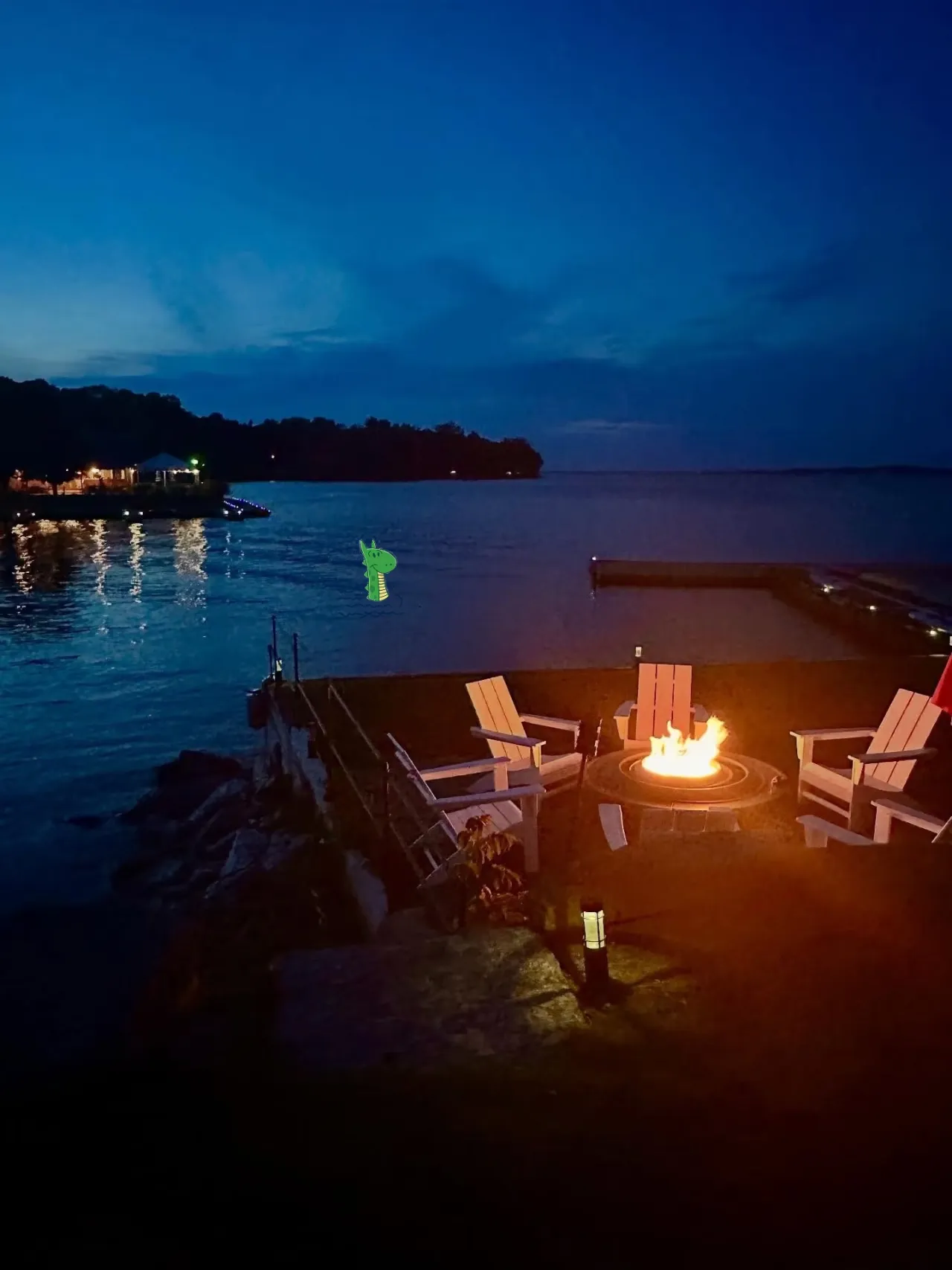 Adirondack chairs surround a lit fire pit on a lakeshore in the evening. A cartoon lake monster peeks out of the water.