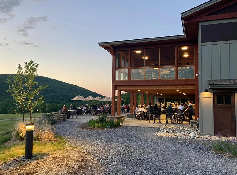 An exterior view of a modern golf course restaurant at sunset, with tables and chairs on a patio.