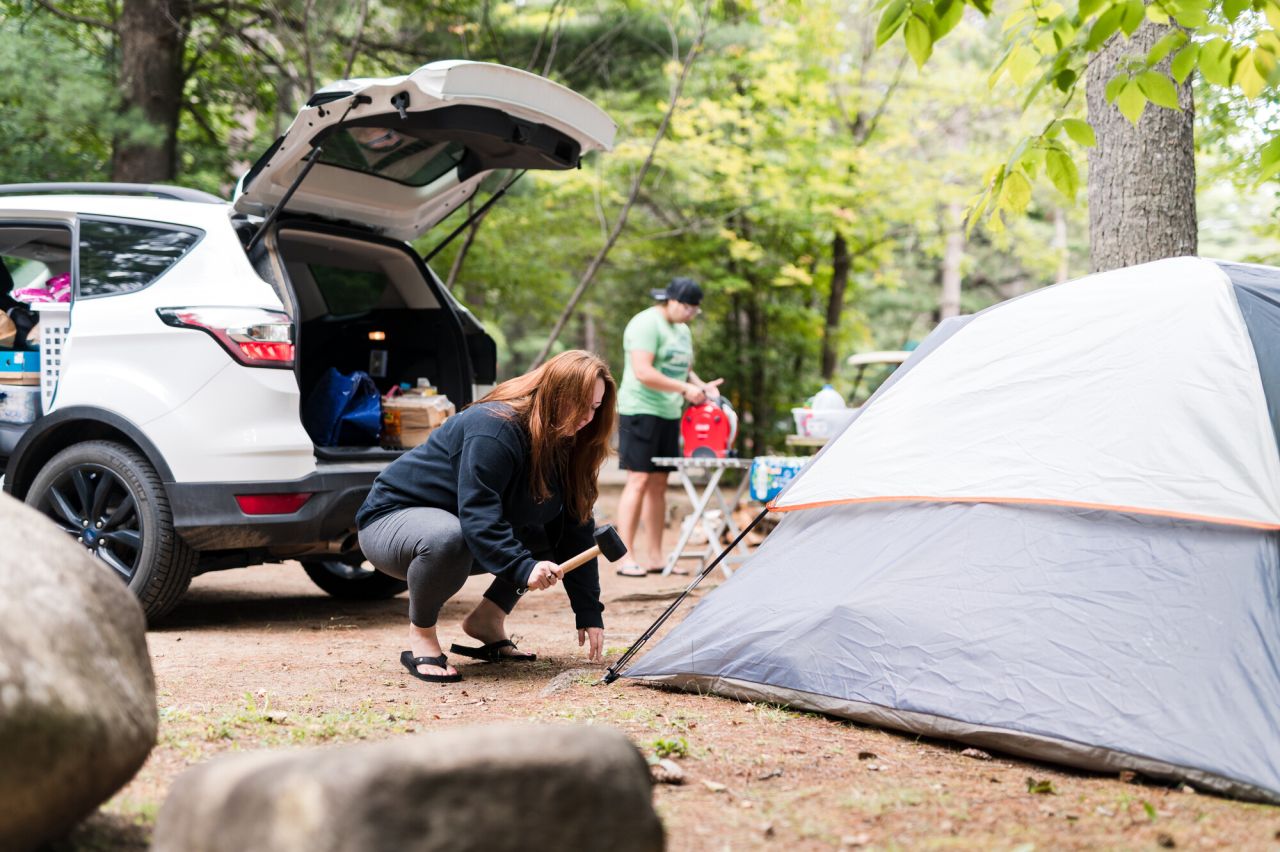 A woman nails a tent corner into the ground with her car trunk open behind her. 