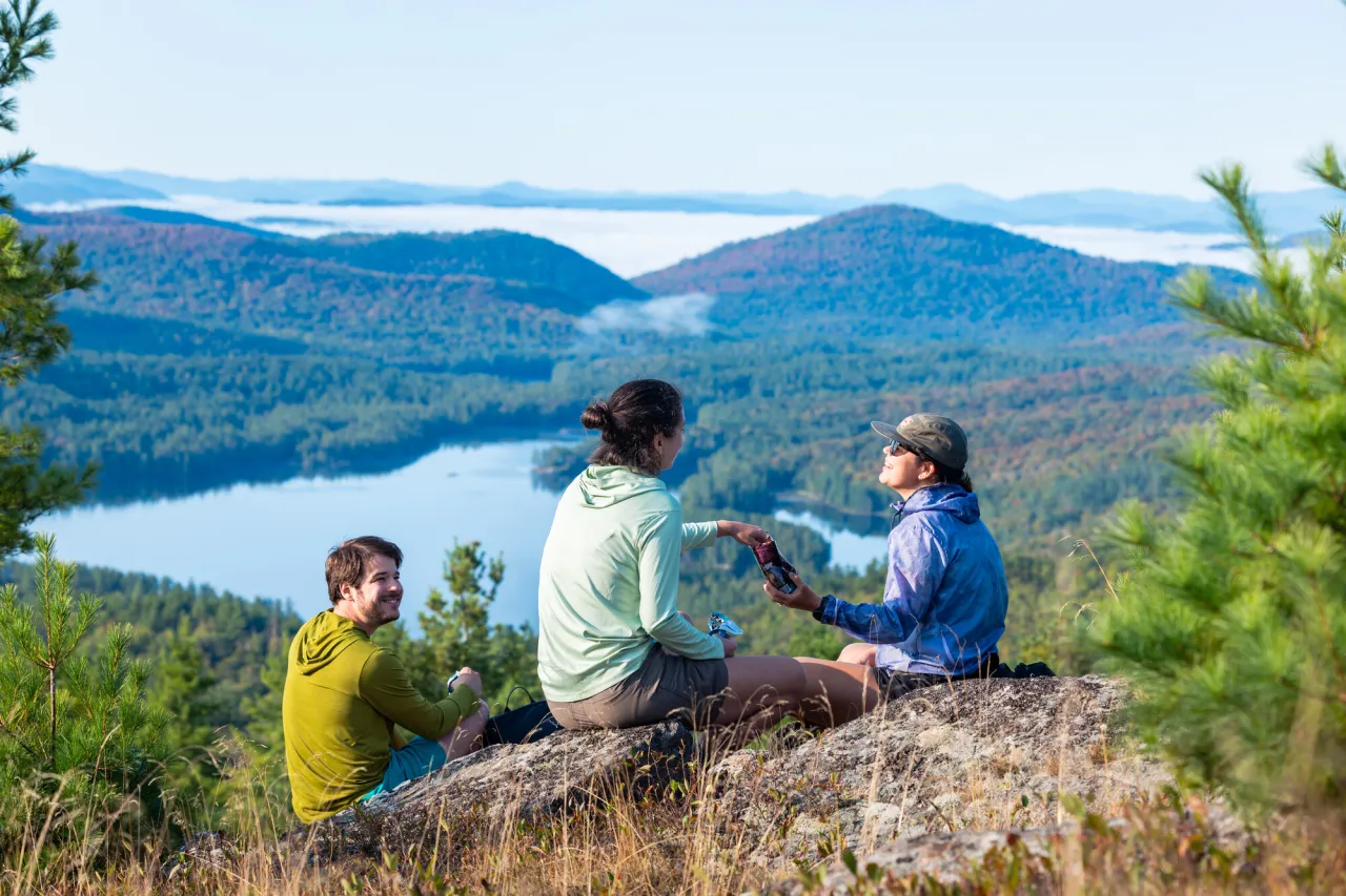 A group of three friends drink water on a mountain top overlooking a lake and mountains below.