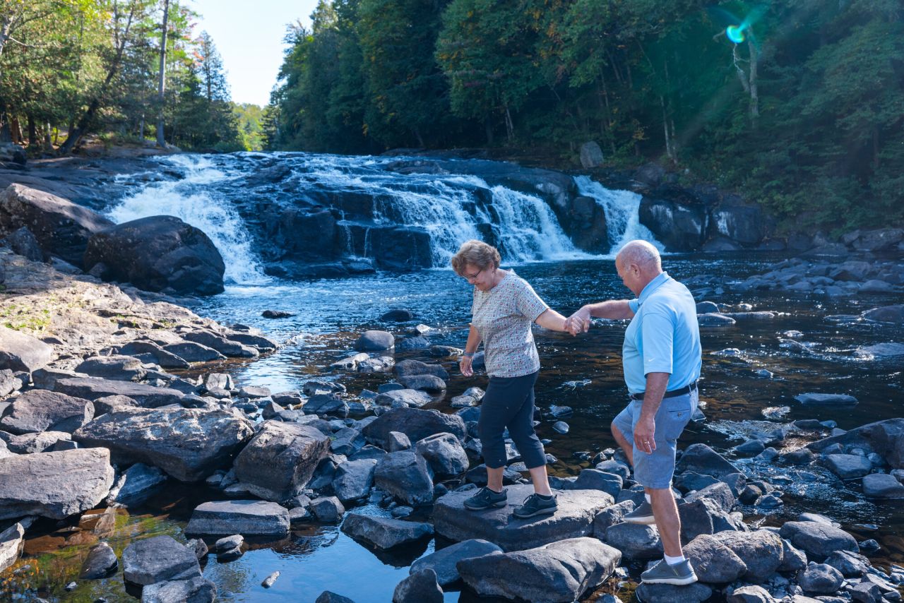 A man and woman walk across a rocky base of a waterfall. 