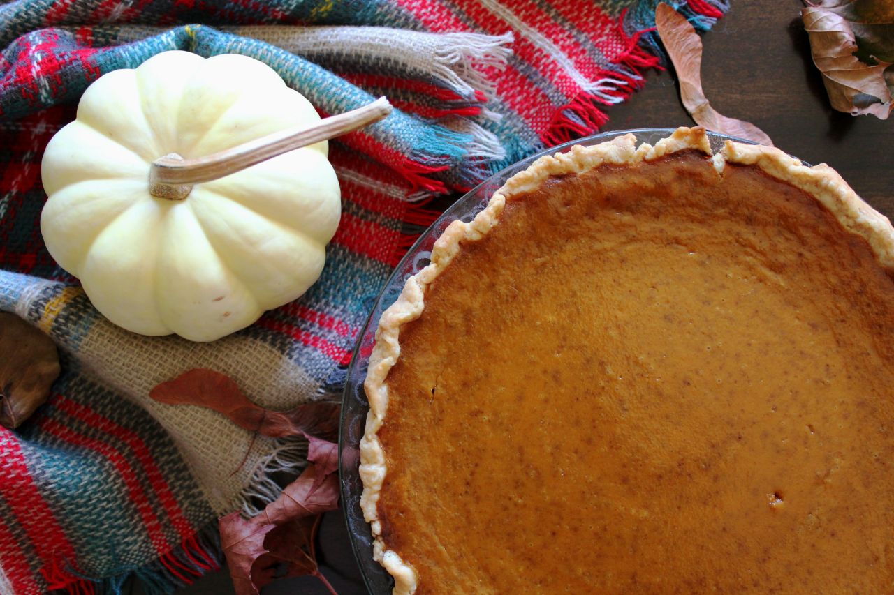 Looking down at a pumpkin pie from above. Next to the pie is a small white gourd and a plaid blanket.
