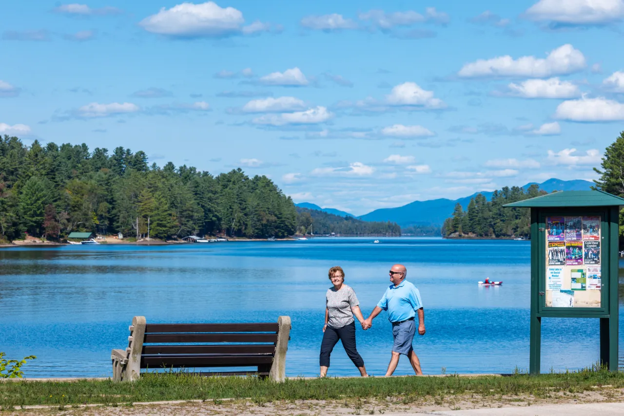 A middle-aged man and woman walk along the shore of a long lake in summer. Tree-covered hills are on either side of the lake.