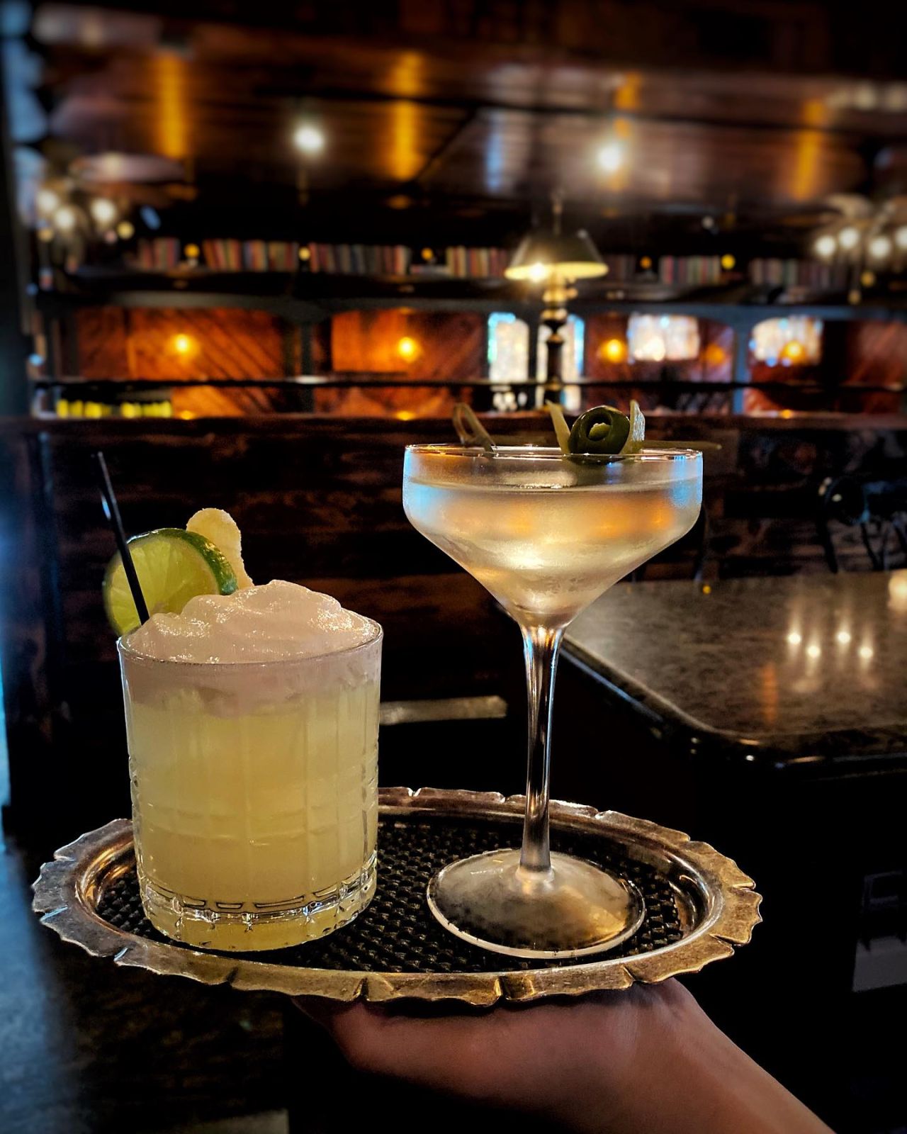 A close up of a tray holding two cocktails, one in a  martini glass. A low lit bar is in the background. 