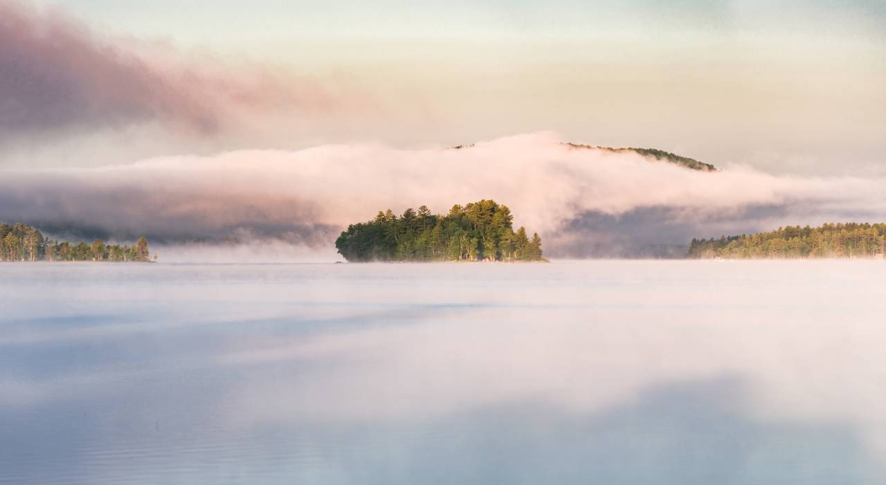 An Adirondack lake is covered by early-morning mist, which surrounds a small, tree-covered island.