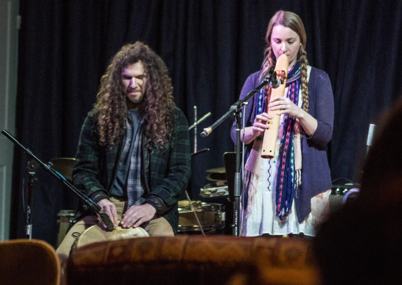 Man with long curly hair plays hand drum next to woman playing a wooden clarinet-style instrument