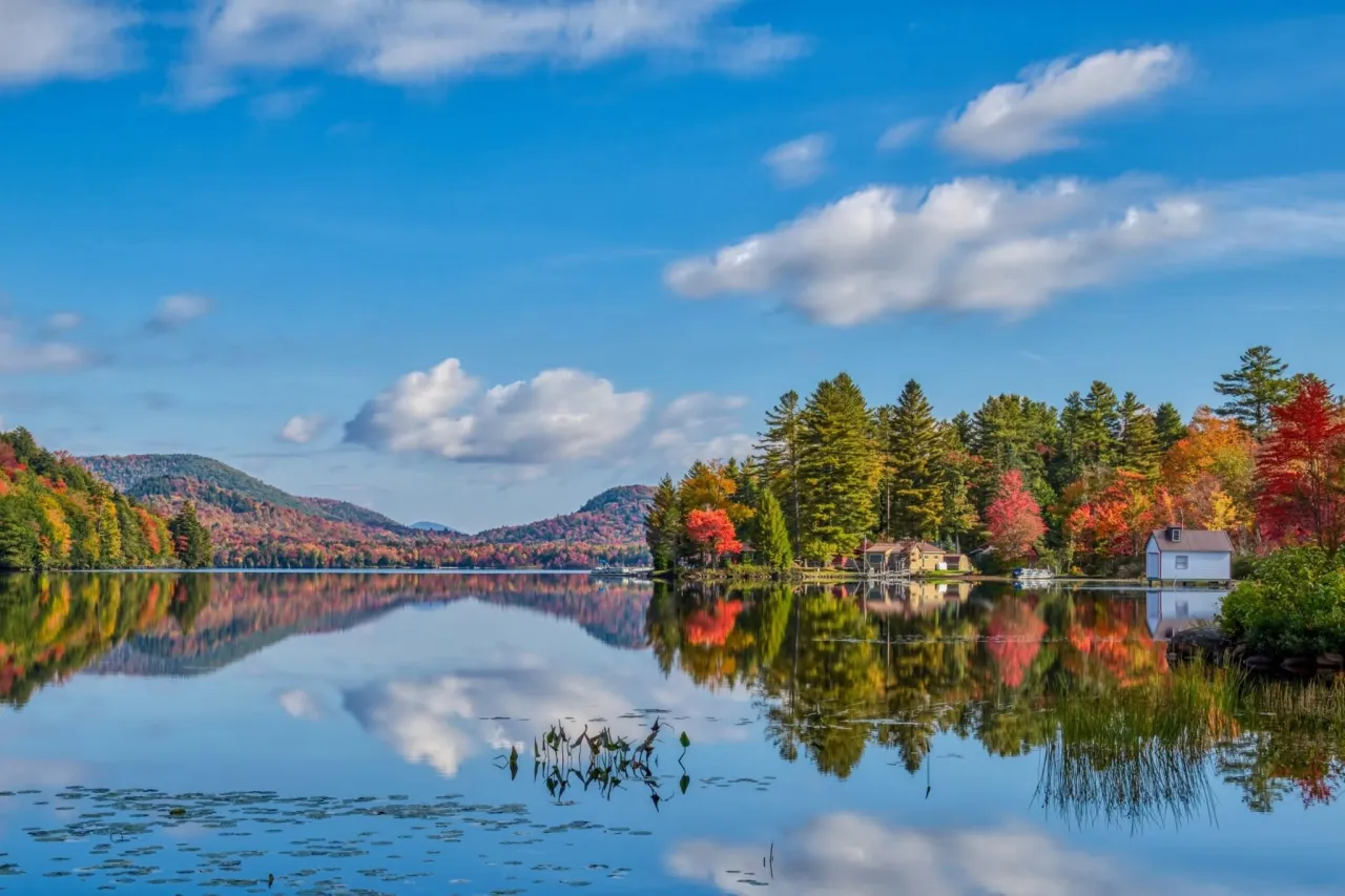 A calm Adirondack lake reflects the blue sky and multi-colored fall foliage. A small cabin is on the shore.